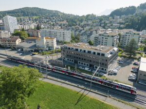 Eine Luftaufnahme eines grossen grauen Gebäudes mit einem Zug im Vordergrund. Im Hintergrund sind weitere Häuser und Bäume zu erkennen