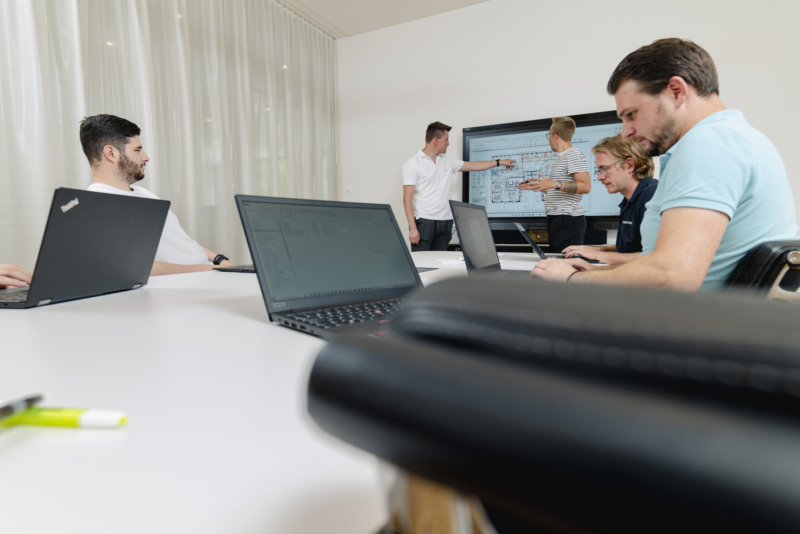 Eine Gruppe von Personen sitzt um einen Tisch herum und arbeitet mit Laptops. Im Hintergrund sind zwei Personen einen Plan am Präsentieren.