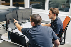 Zwei Personen sitzen nebeneinander am Arbeitsplatz mit zwei Bildschirmen und besprechen etwas was auf dem Bildschirm zu sehen ist. Die eine Person zeigt mit dem Finger auf einen der beiden Bildschirme.